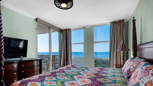 A bedroom with a large bed, a TV on a dresser, and a balcony with a view of the ocean. - Emerald Dolphin Unit 510 (Pensacola Beach)