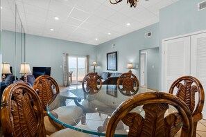 A dining area with a glass-top table surrounded by wooden chairs, a television, and a view of the outdoors. - Sans Souci Unit 104 (Pensacola Beach)