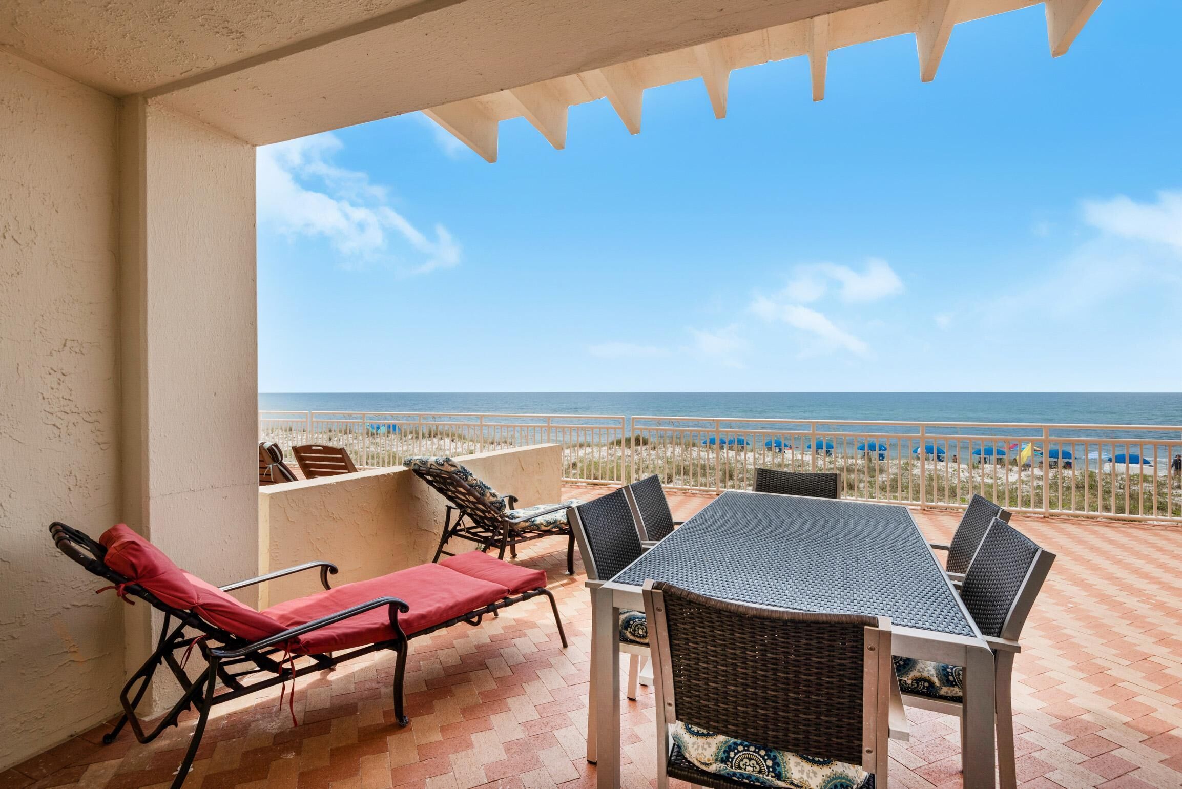 A terrace with a table and chairs, a red lounge chair, and a view of the beach.
