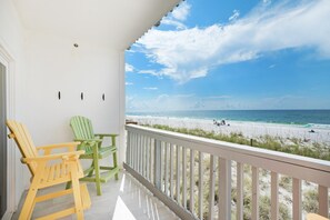 A balcony with two Adirondack chairs, a white railing, and a view of a beach with people and waves. - Villas on the Gulf Unit M5 (Pensacola Beach)