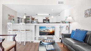 A modern living room with a flat-screen TV, a leather sofa, and a dining area with white chairs. - Starboard Village 424- Beach Haven (Pensacola Beach)