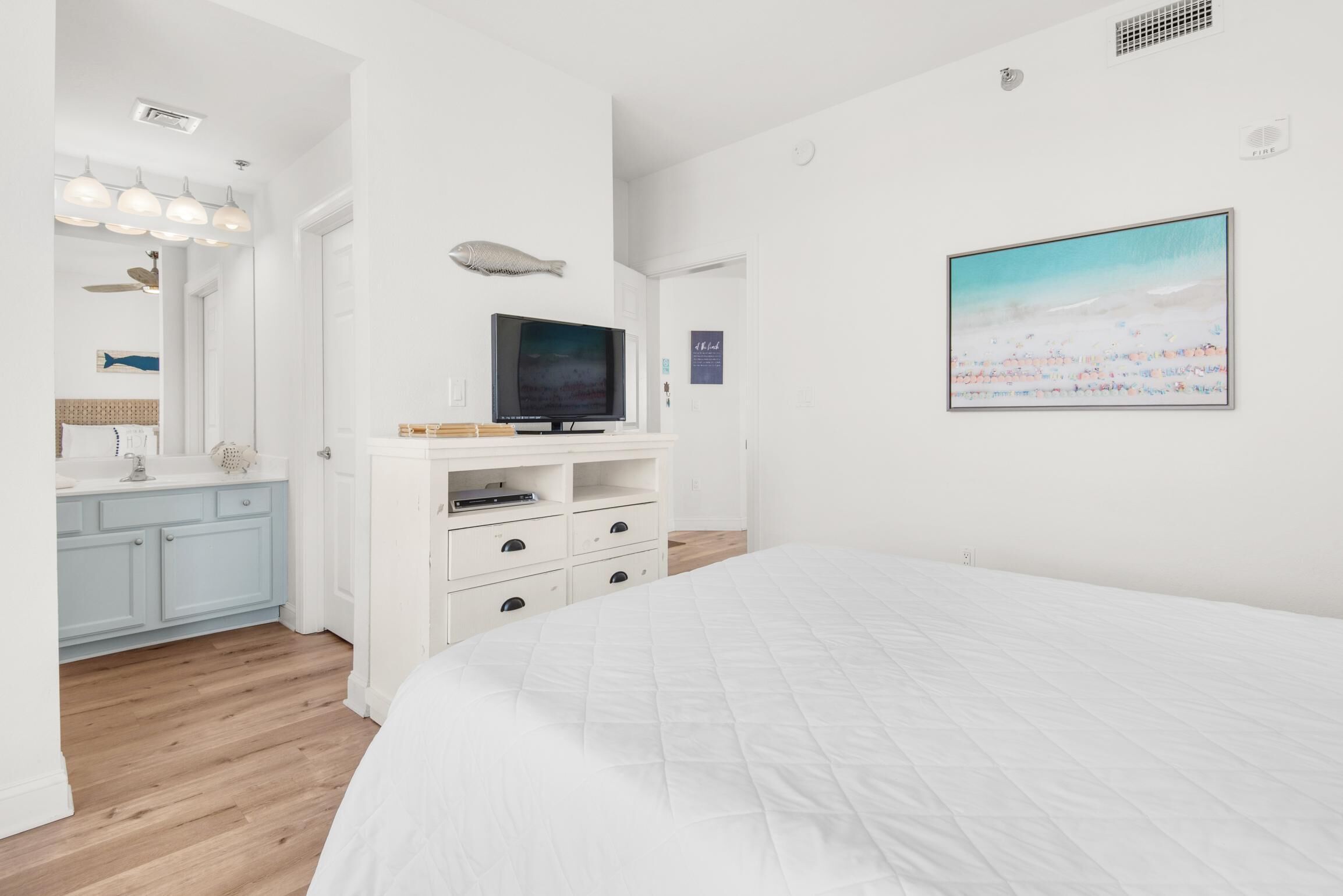 Une chambre à coucher avec un grand lit, un téléviseur posé sur un meuble blanc, un tableau accroché au mur, et une salle de bain avec un meuble bleu et une décoration sur le thème des poissons.