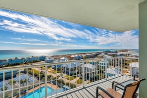 A balcony with a view of a beach and ocean, a pool, and several buildings with blue roofs. - Tristan Towers Unit 9B (Pensacola Beach)
