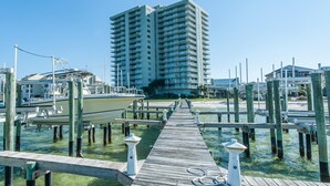 A wooden pier leading to a marina with boats docked, a tall building in the background, and clear skies. - Tristan Towers Unit 9B (Pensacola Beach)