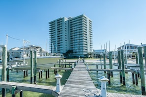 A wooden pier leading to a marina with boats docked, a tall building in the background, and clear skies. - Tristan Towers Unit 9B (Pensacola Beach)