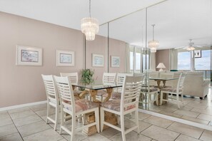 A dining area with a glass-top table surrounded by white chairs with cushioned seats. There are framed artworks on the wall, a chandelier hanging above, and a large window providing natural light.