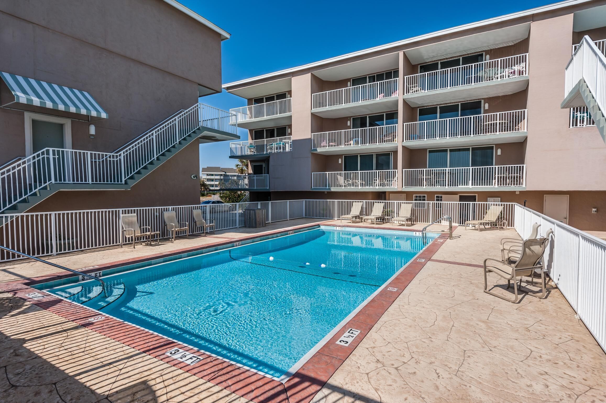 A swimming pool with a sun deck, surrounded by a multi-story building with balconies and a stairway.