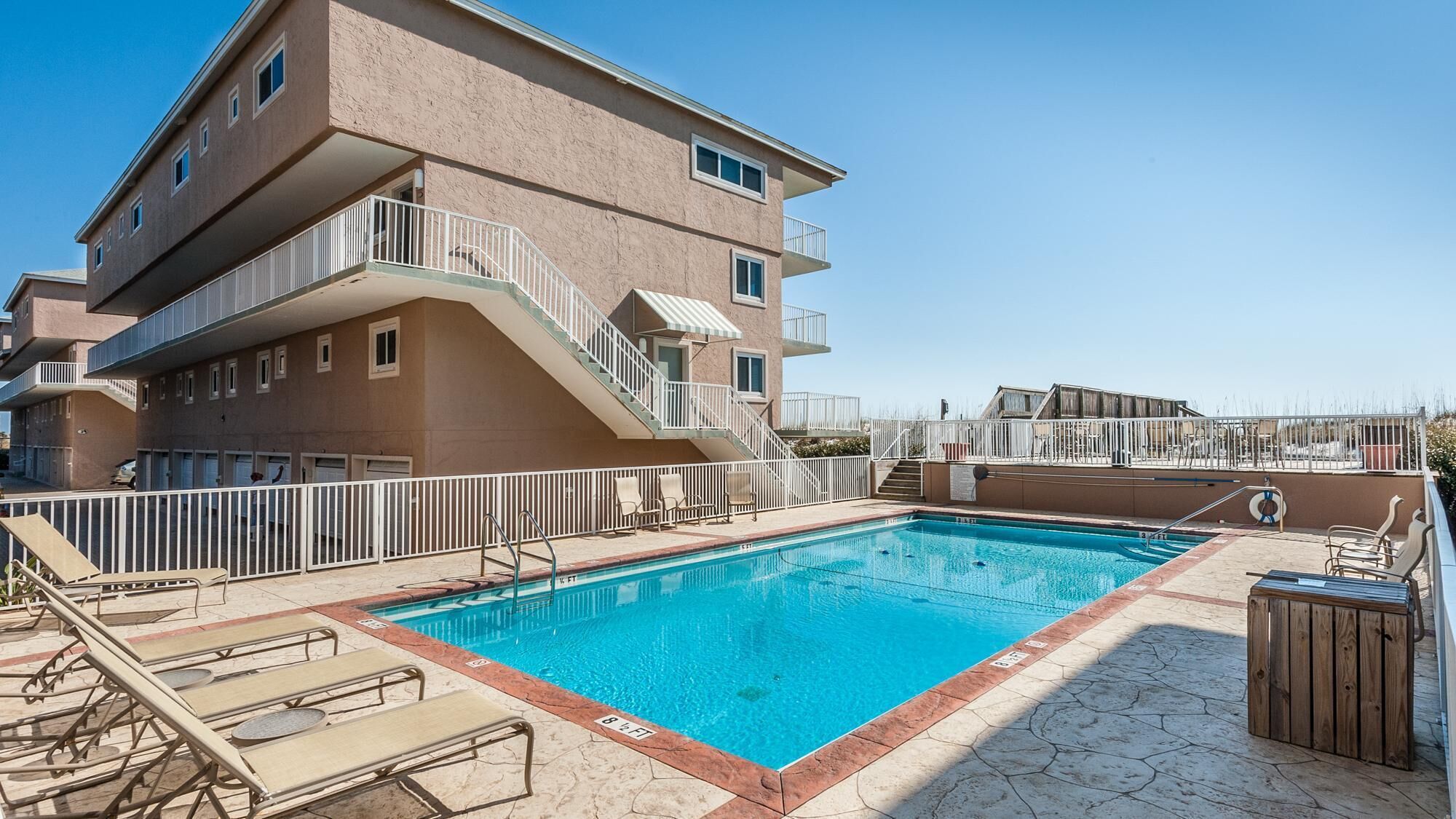 A swimming pool with sun loungers and a wooden deck, surrounded by a multi-story building with balconies.