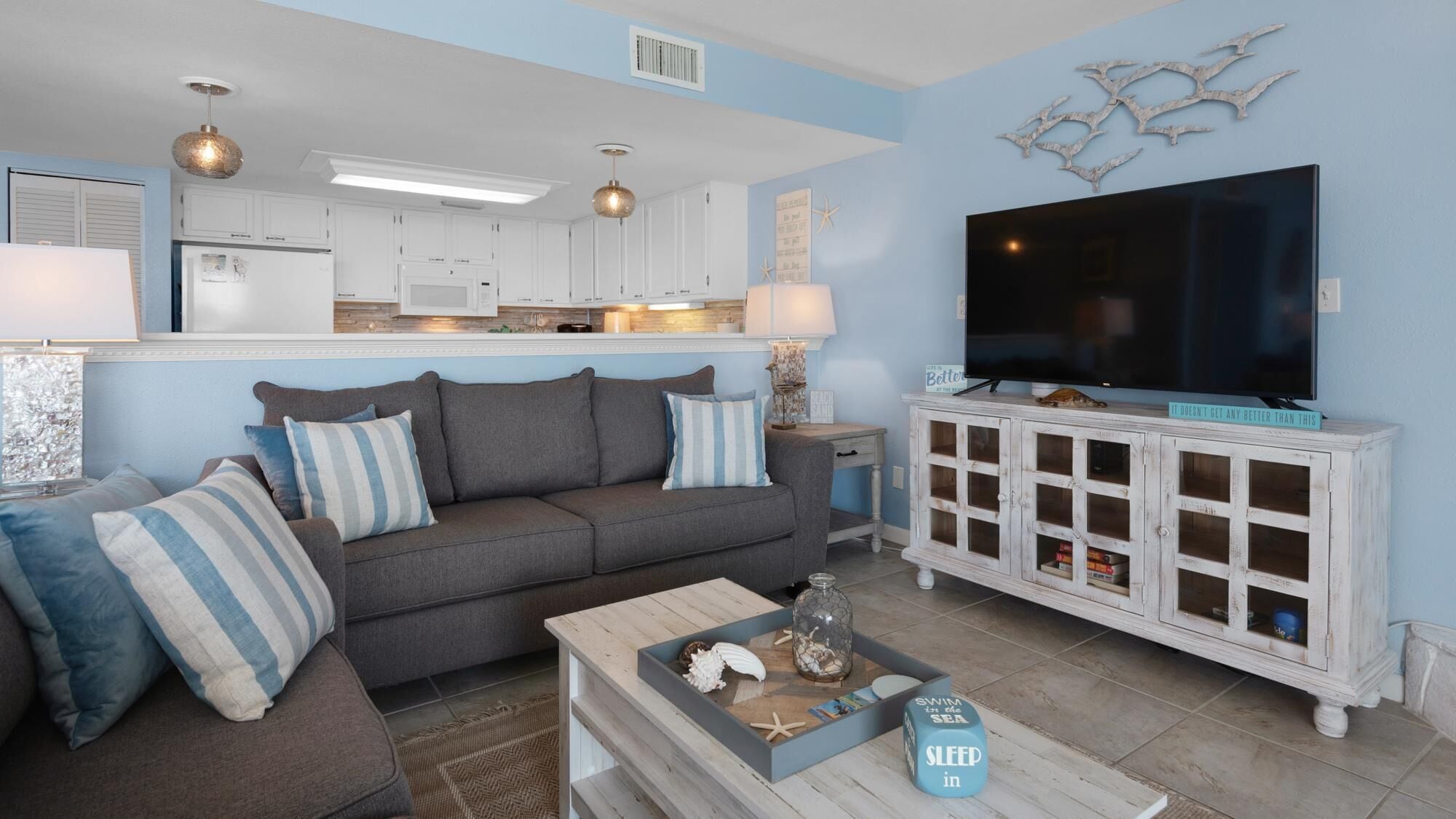 A living room with a grey sofa, striped pillows, a coffee table with decorative items, a TV on a white cabinet, and a kitchen in the background.