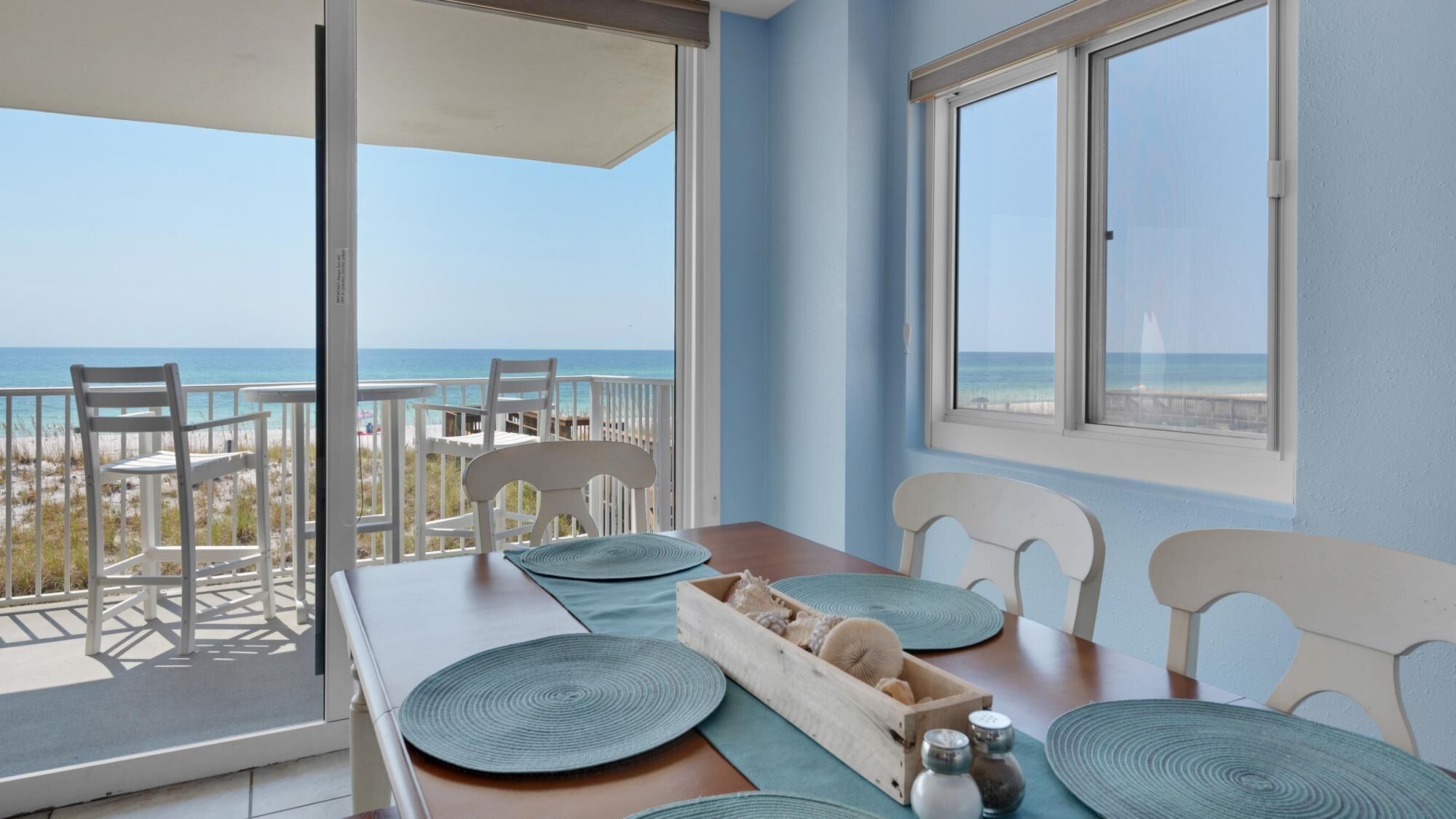 A balcony with a view of the beach and ocean, a wooden table with blue placemats, and white chairs.