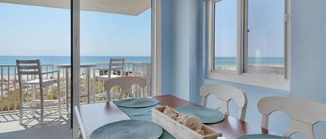 A balcony with a view of the beach and ocean, a wooden table with blue placemats, and white chairs.