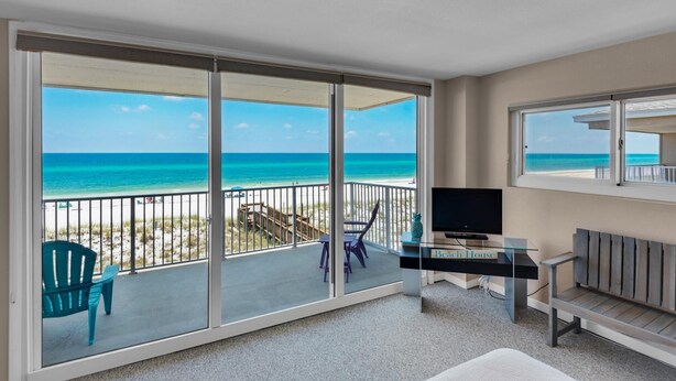 Un balcon avec une porte vitrée donnant sur la plage, un téléviseur posé sur une table en verre et un coin salon avec un banc et des chaises.