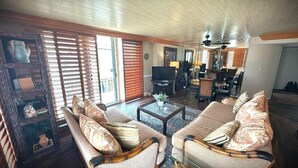 A spacious living room with a large sofa, a coffee table, and a television. There is a dining area with chairs and a table in the background. - Tristan Towers Unit 1B (Pensacola Beach)
