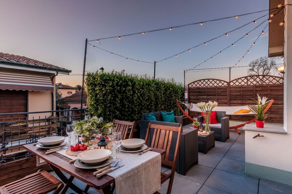 An outdoor patio with a dining table set, outdoor seating, string lights, and a hedge.
