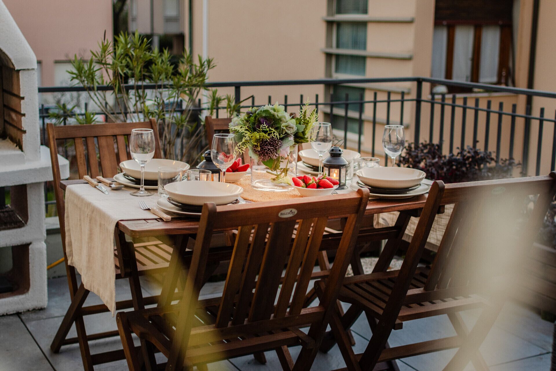 A dining table set outdoors with wooden chairs, plates, glasses, and a centerpiece of flowers and strawberries.