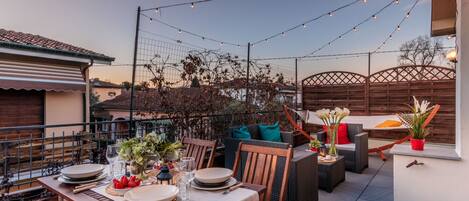 Une terrasse extérieure avec une table à manger pour quatre personnes, entourée de meubles en osier et illuminée de guirlandes lumineuses.