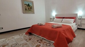 A neatly made bed with a red bedspread and white pillows, a small bedside table with a lamp, and a patterned rug on the floor. - Housestation suite H (Palermo)