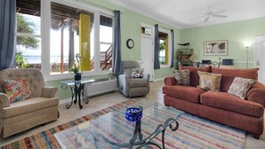 Un salon avec un canapé beige, un canapé rouge, une table basse en verre et un tapis à motifs. On y trouve un fauteuil avec un coussin, une petite table avec un vase et une horloge murale.
