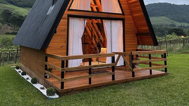 A wooden cabin with a triangular roof, a balcony, and a view of a vineyard in the background. - Refúgio do Vale Cabanas Exclusivas (Bento Gonçalves)