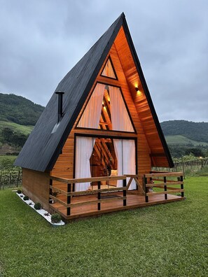 A wooden cabin with a triangular roof, a balcony, and a view of a vineyard in the background. - Refúgio do Vale Cabanas Exclusivas (Bento Gonçalves)