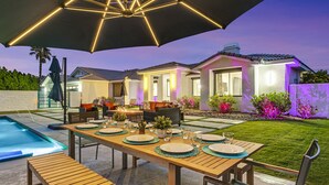 A well-lit outdoor dining area with a pool, a covered table, and a patio with a view of a house. - Rising Sun House: Pool, Spa, GameRoom, PetFriendly! (Palm Springs)