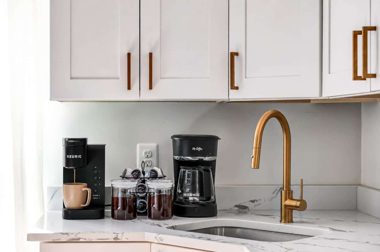 A kitchen with white cabinets, a coffee maker, a coffee pot, a coffee grinder, and a gold faucet.