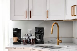 A kitchen with white cabinets, a coffee maker, a coffee pot, a coffee grinder, and a gold faucet.