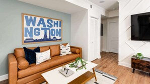 Ein Wohnzimmer mit einem braunen Ledersofa, einem weißen Couchtisch, einem gerahmten 'WASHINGTON'-Schild und einem Fernseher an der Wand.