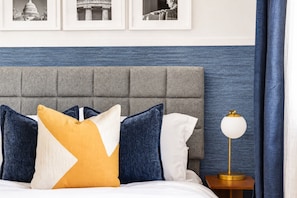 Ein Bett mit grauem Polsterkopfteil, ein Nachttisch mit goldener Lampe und drei gerahmte Bilder an der Wand.