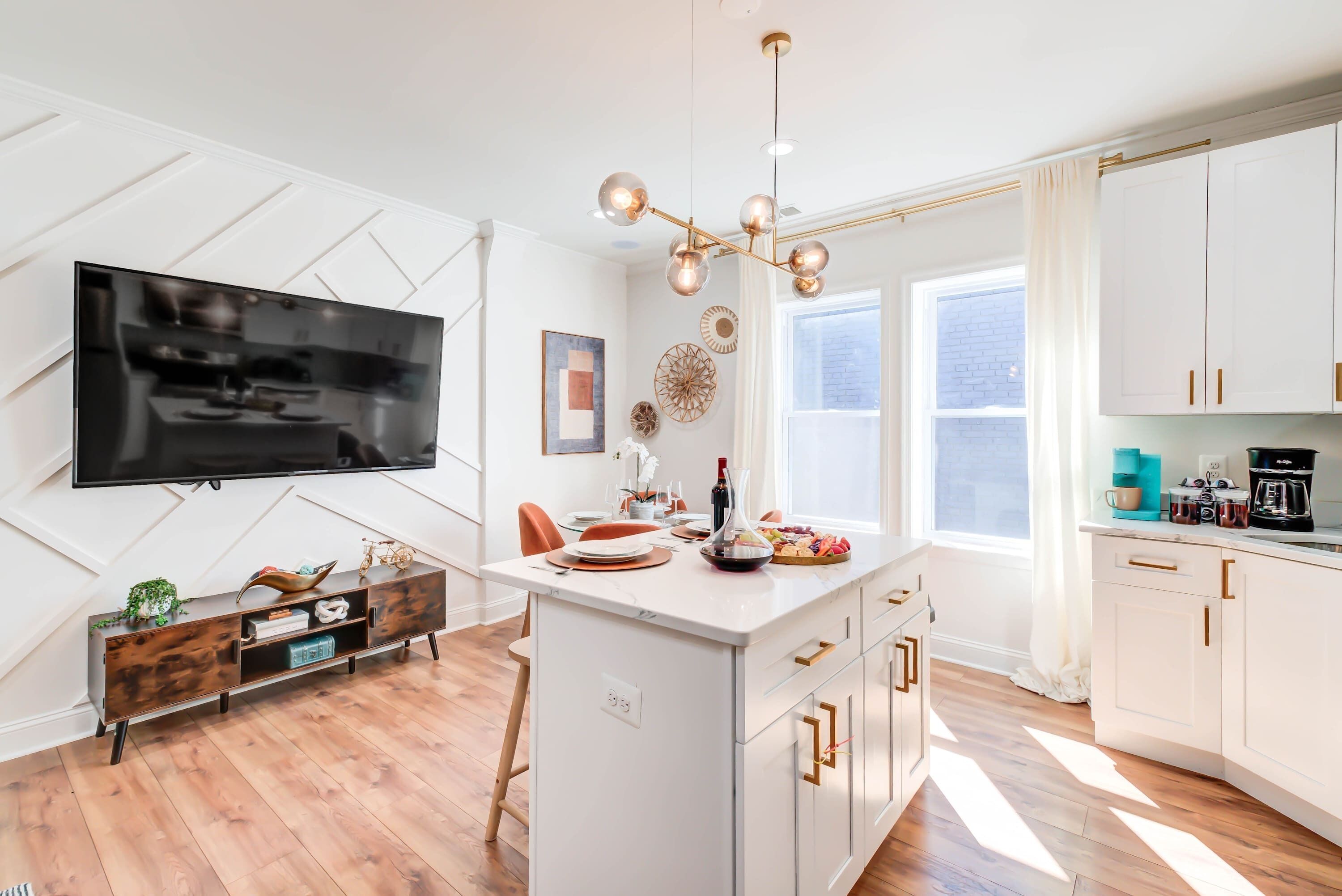 Een moderne keuken met een centraal eiland, witte kasten, een flatscreen televisie en een hanglamp.
