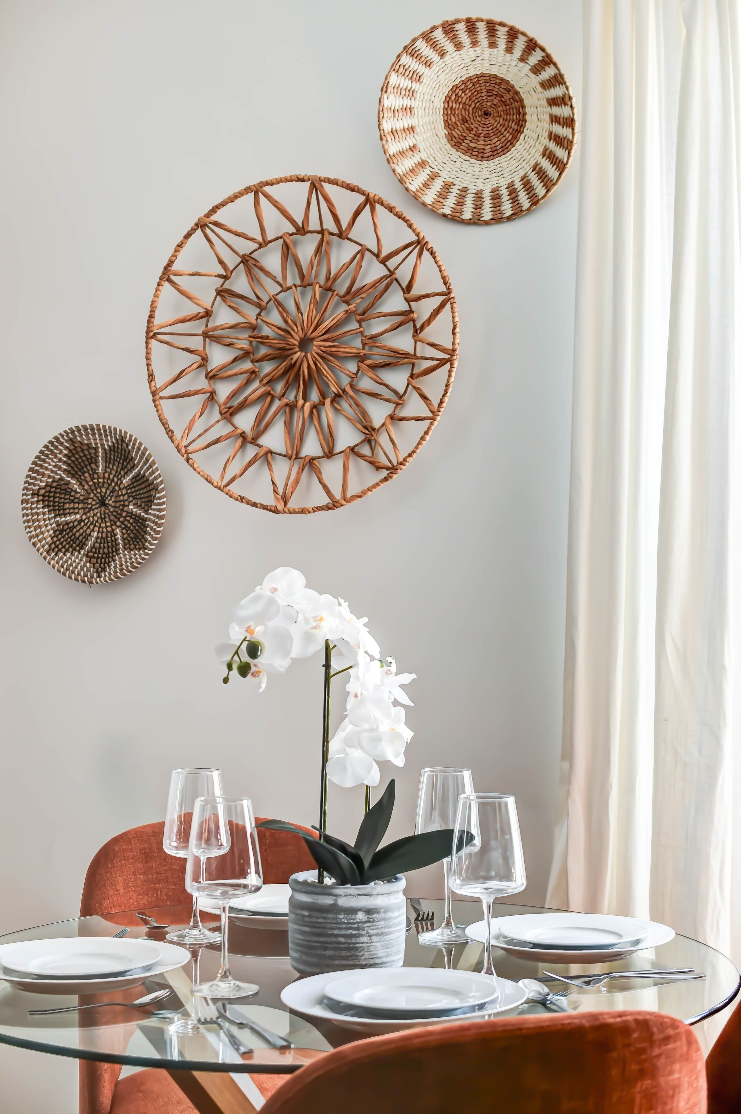 A dining area with a glass table, white plates, and wine glasses, surrounded by orange chairs and a white orchid plant.
