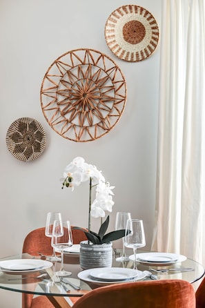 A dining area with a glass table, white plates, and wine glasses, surrounded by orange chairs and a white orchid plant.