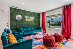 A modern living room with a teal sofa, red ottomans, a disco ball, and a neon sign on the wall.