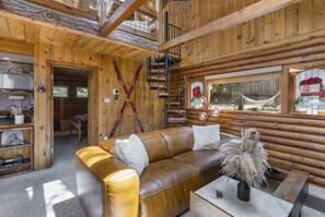 Interior de una cabaña de madera con sofá de cuero, zona de comedor, escalera y una hamaca junto a una ventana.