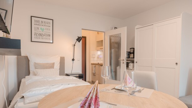 Habitación de hotel con una cama bien hecha, una mesa redonda con un mantel individual floral y una cita enmarcada en la pared.