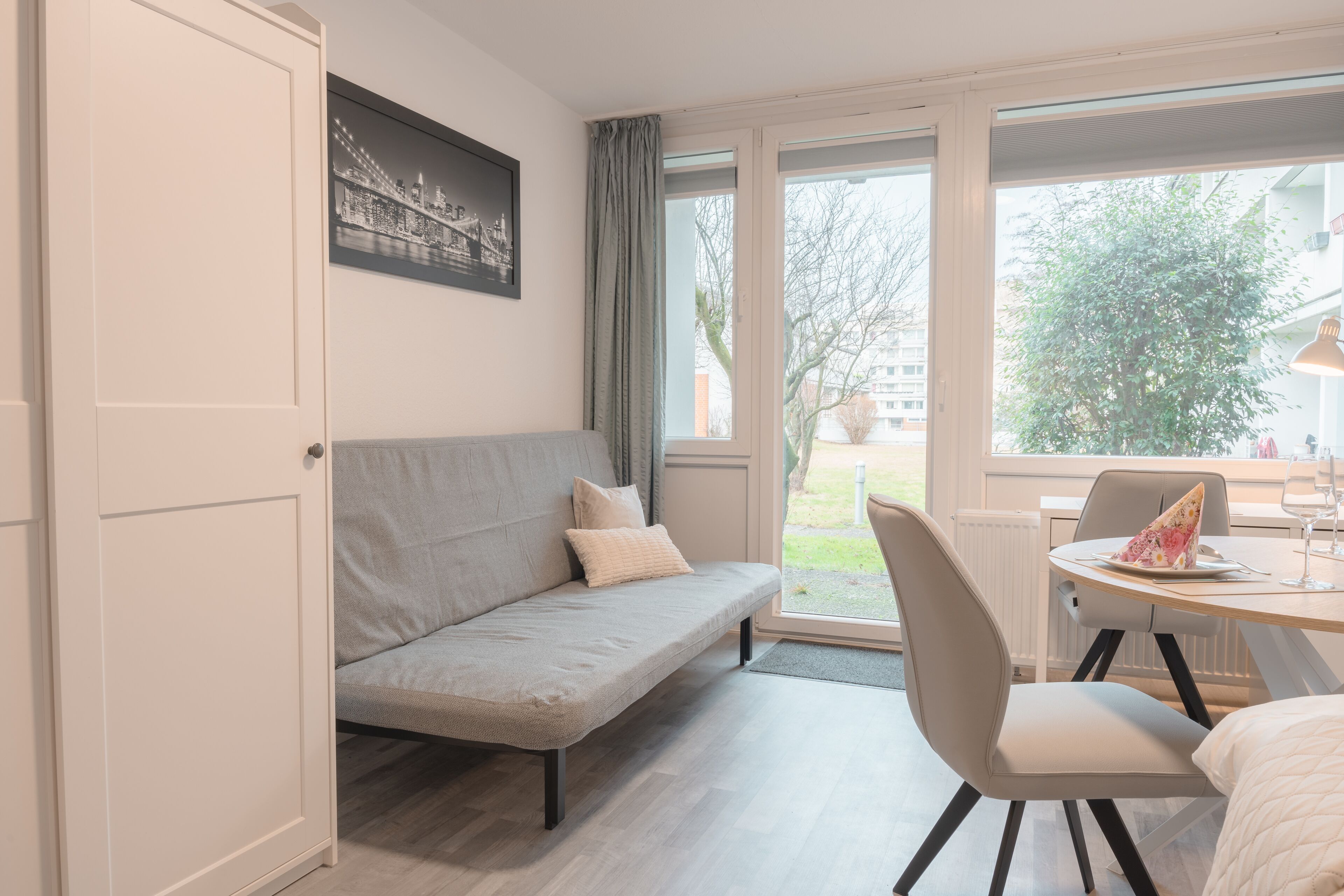A modern room with a grey sofa, a dining table with chairs, and a window with a view of the outdoors.