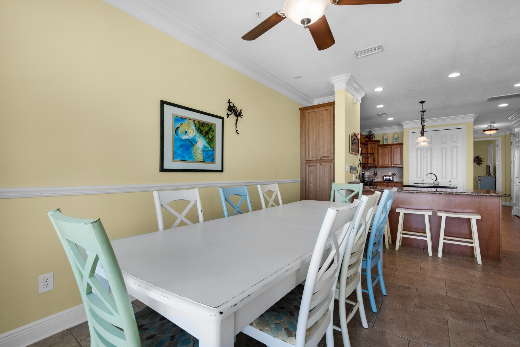 A dining area with a white table and chairs, a ceiling fan, recessed lighting, and a kitchen in the background.