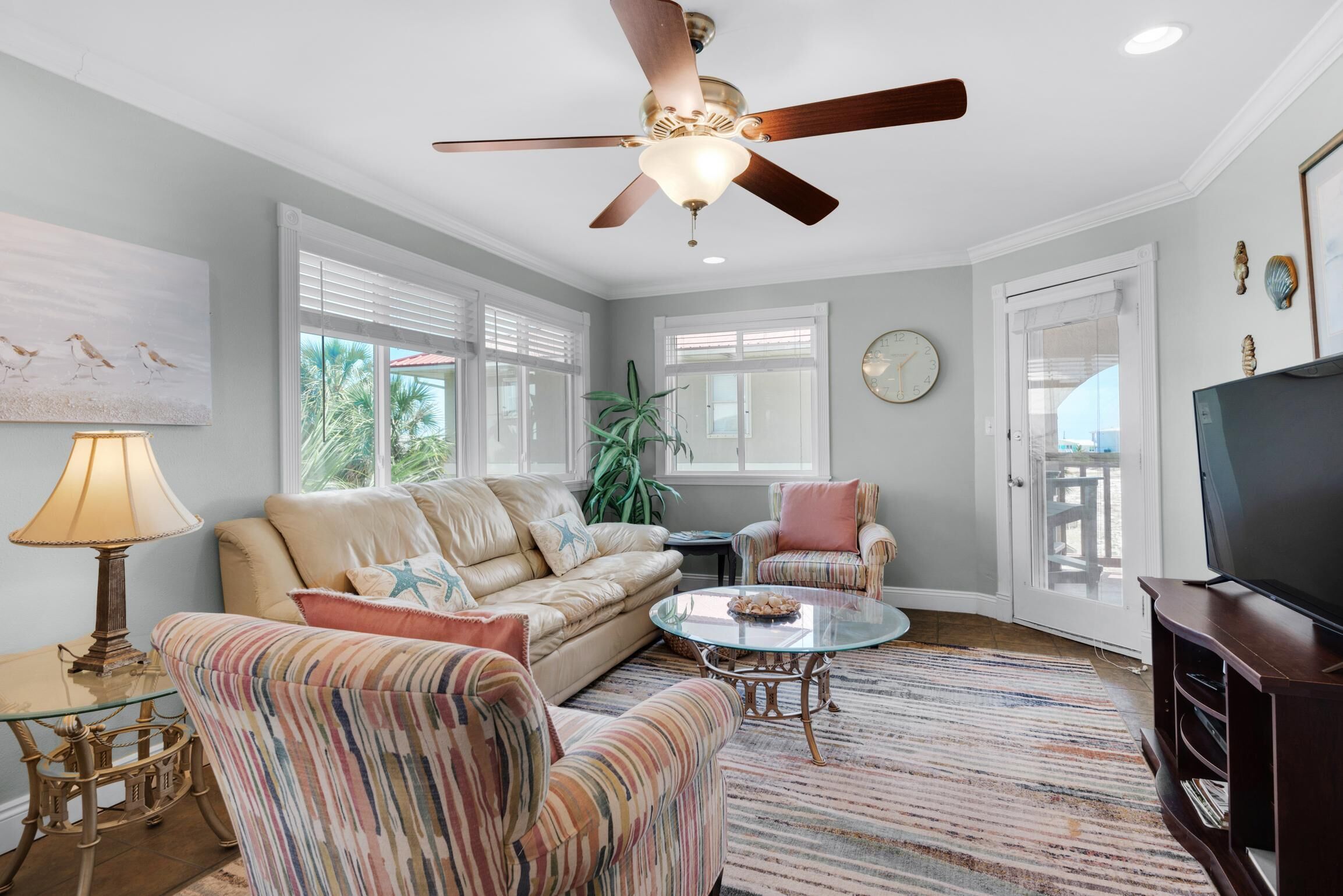 Un salon avec un ventilateur de plafond, un canapé, un fauteuil à rayures, une table basse en verre et un téléviseur.