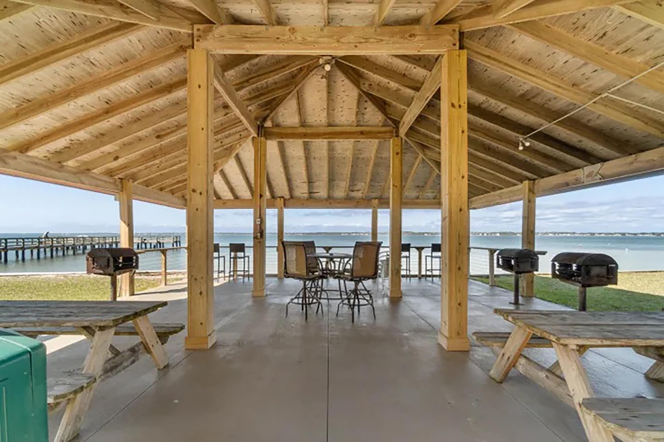 A covered pavilion with picnic tables and chairs, a view of the water, and a pier in the background.