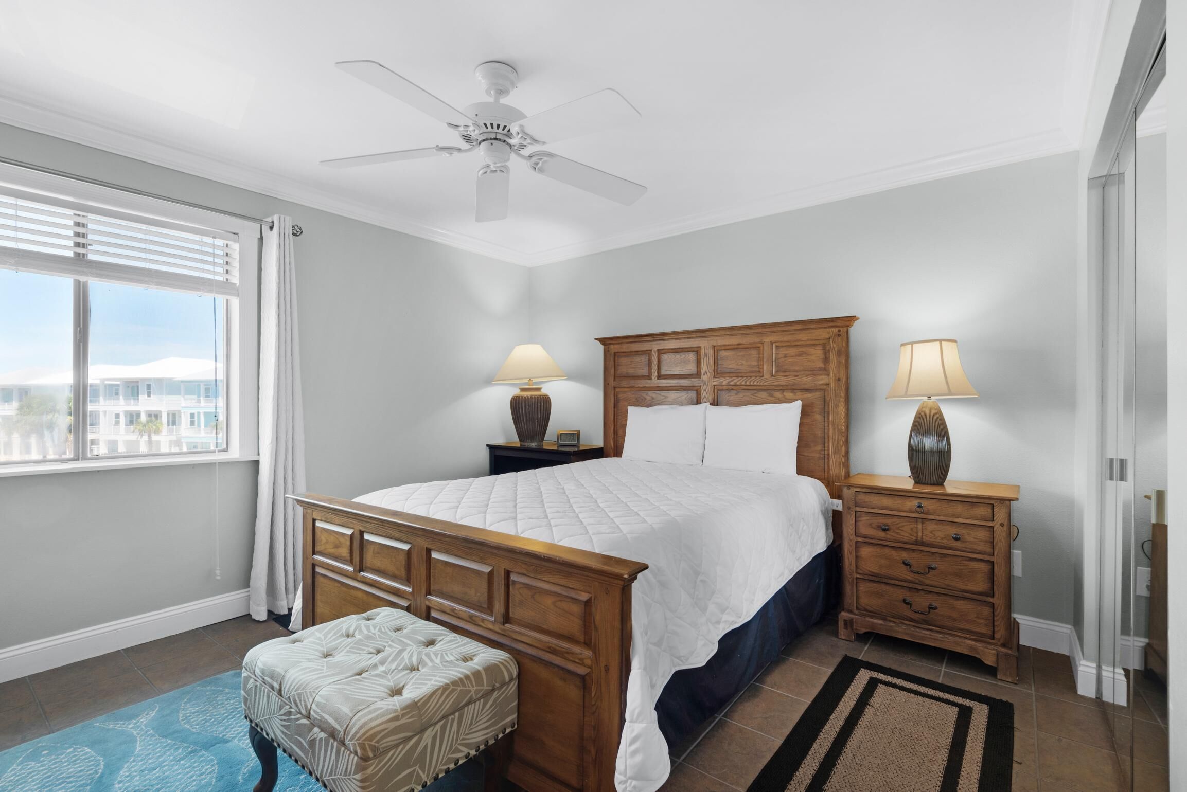 Une chambre à coucher avec un lit en bois, deux tables de chevet avec des lampes, un ventilateur de plafond, une fenêtre avec des stores et une vue sur des immebles.