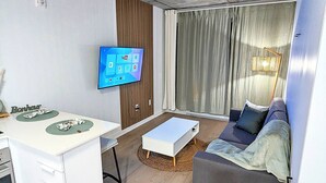Un salon moderne comprenant un canapé, une table basse, un téléviseur à écran plat fixé au mur et un coin cuisine avec un meuble blanc et un petit îlot.