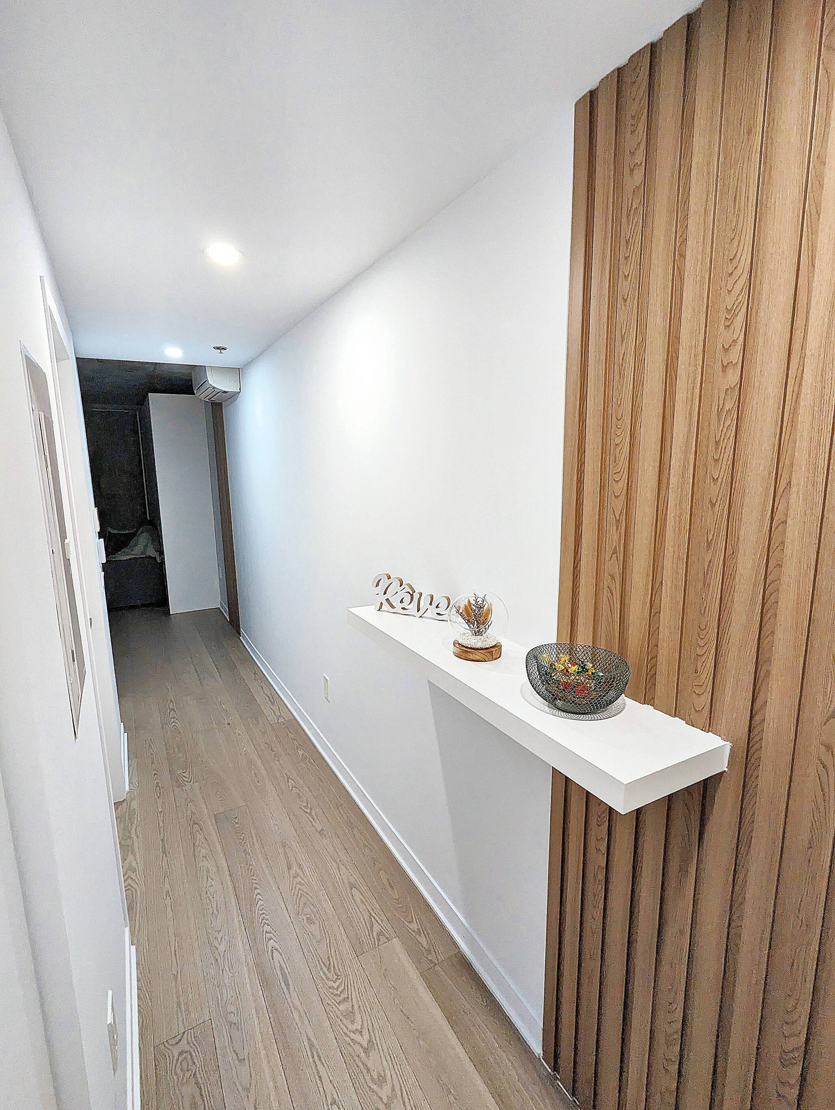 A hallway with a wooden floor, white walls, and a wooden panel on the right. A white shelf with decorative items is mounted on the wall.