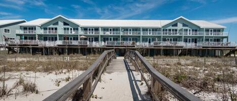 Un hotel frente a la playa con un paseo marítimo de madera que lleva a la entrada, con varios balcones y ventanas amplias.