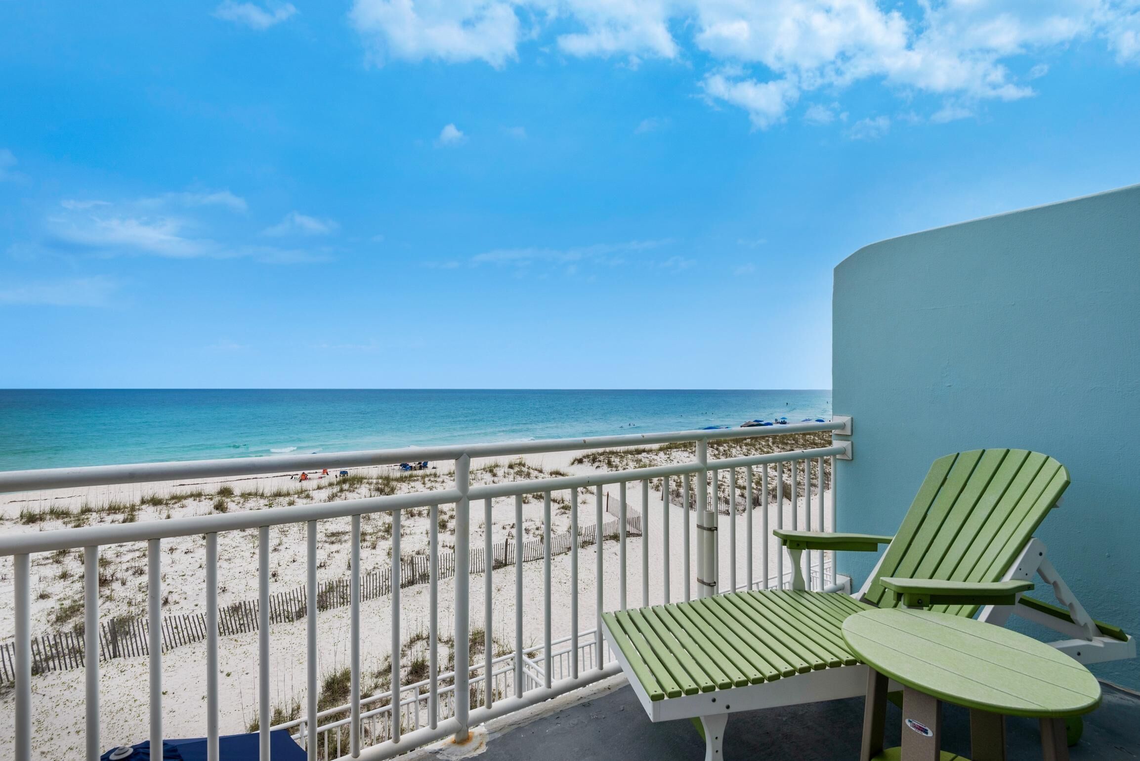 A balcony with a view of a beach, a white railing, and two green Adirondack chairs.