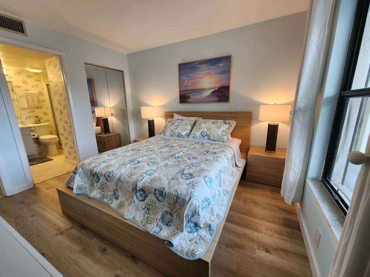 Un dormitorio con cama, lámparas de noche, un cuadro en la pared y una ventana con cortinas.