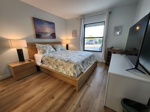 Ein modernes Schlafzimmer mit einem hölzernen Bett, Nachttischen, einem Fernseher und einem Fenster mit Blick auf geparkte Autos.
