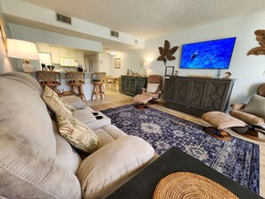 Una sala de estar con sofá, sillas, televisión y una cocina al fondo.