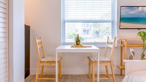 Une pièce avec un bureau blanc, deux chaises en bois, une petite table avec une plante et un lit avec une tête de lit blanche.