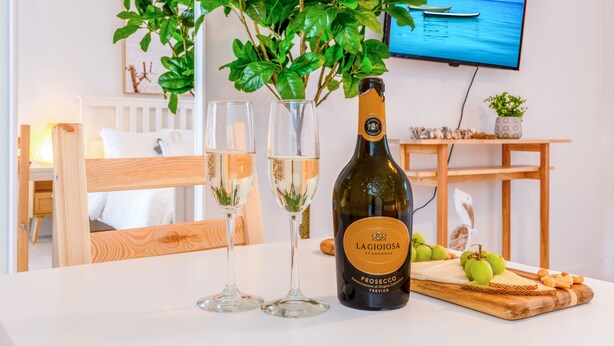 Une bouteille de Prosecco Lagioiosa, deux verres, une planche à découper en bois avec des raisins et des crackers, une plante en pot et un téléviseur diffusant une scène côtière.