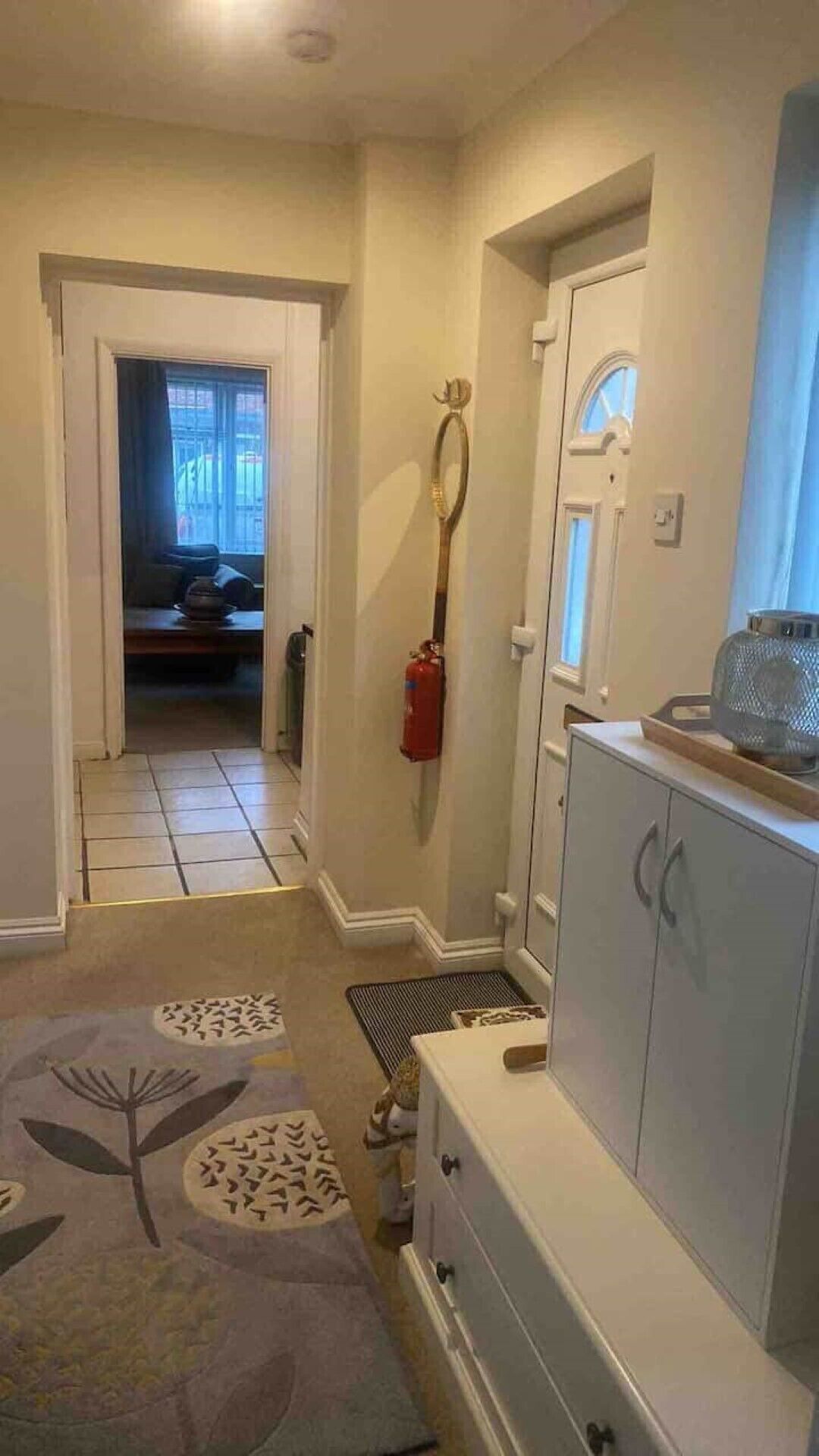 A hallway with a tiled floor, a patterned rug, a fire extinguisher mounted on the wall, and a cabinet with drawers.
